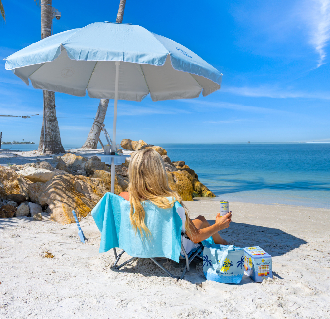 Woman sitting in a beach chair under a blue umbrella looking at the ocean.