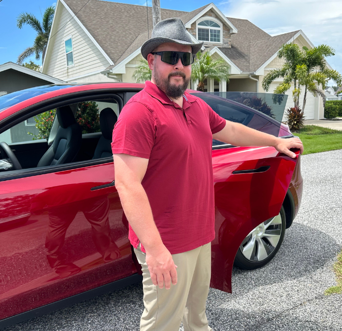 Man standing next to a red car parked in a coastal residential neighborhood.