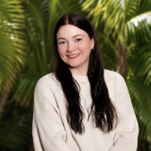 Portrait of a smiling female employee with long brown hair wearing a white top.