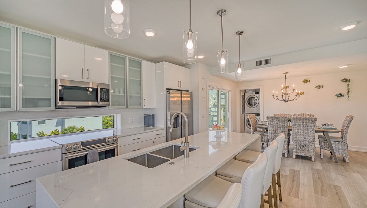 A bright and modern open-concept kitchen at a Cottages on the Key vacation rental, featuring a large quartz island with a double sink, high-end stainless steel appliances, and a chic dining area with wicker chairs and a coastal-inspired chandelier.