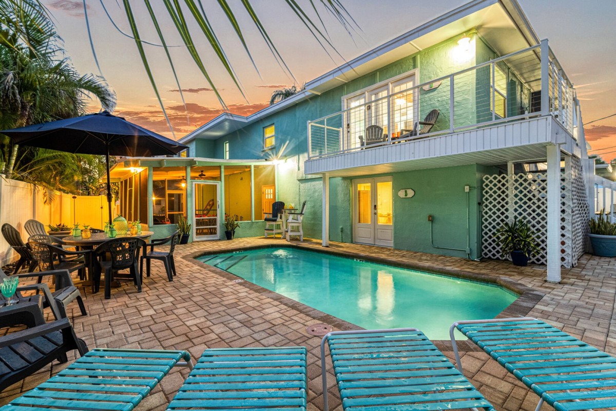 A beautiful two-story teal vacation home managed by Cottages on the Key, showcasing a lit private pool, outdoor dining area, and upper-level balcony against a vibrant Florida sunset, creating a serene evening atmosphere for guests.