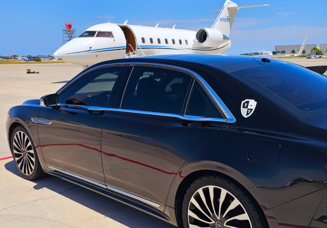 Black luxury sedan on a runway, with a white private jet in the background and a T logo on the car's rear window.